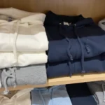 A neatly folded pile of cozy sweatshirts in neutral tones on a wooden shelf