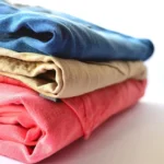 A neatly folded stack of colorful t-shirts on a wooden shelf